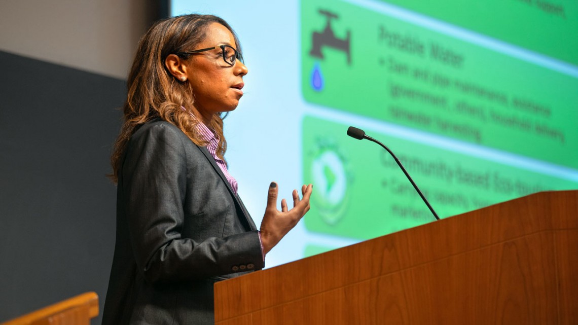 Professor Shorna Allred stands at a podium with a presentation on indigenous sustainability research visible in the background