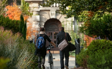 Two students wearing jackets walk a bike through campus greenery in the Fall